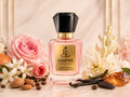 A bottle of Royal Essence Vampire inspired by Good Girl perfume, featuring a pink liquid, a gold label, and a faceted black cap. The bottle is beautifully styled with pink roses, white flowers, vanilla beans, almonds, and a piece of amber on a marble surface with a soft beige background.